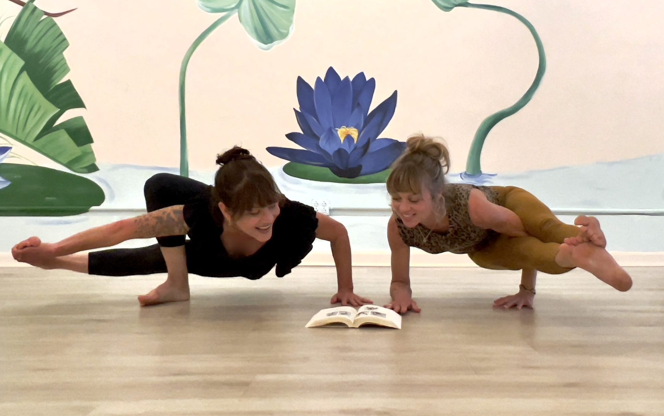 Jasmine and Kate in an arm balance pose looking at a book.